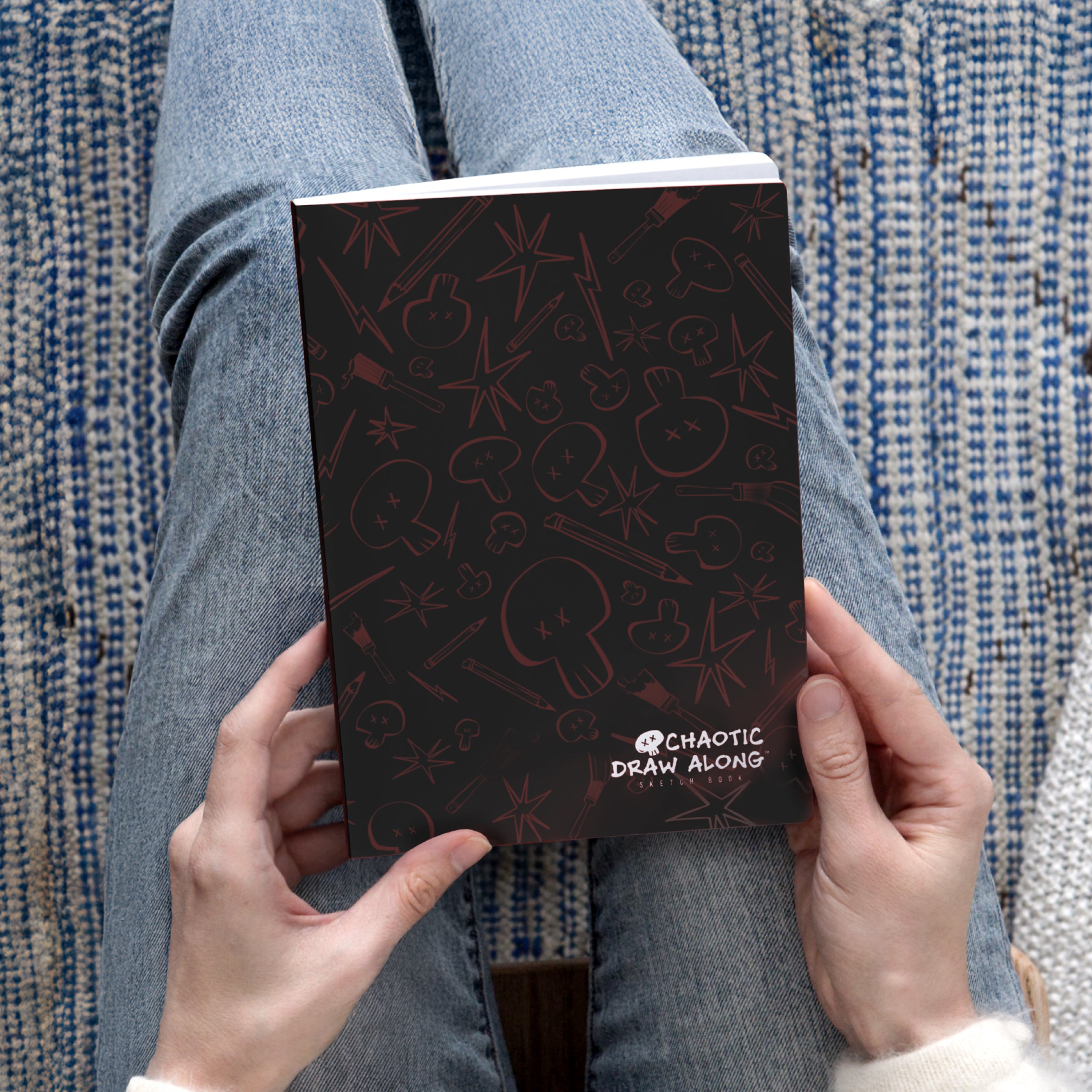 Person holding the black Chaotic Draw Along Sketchbook while seated on a blue patterned rug.