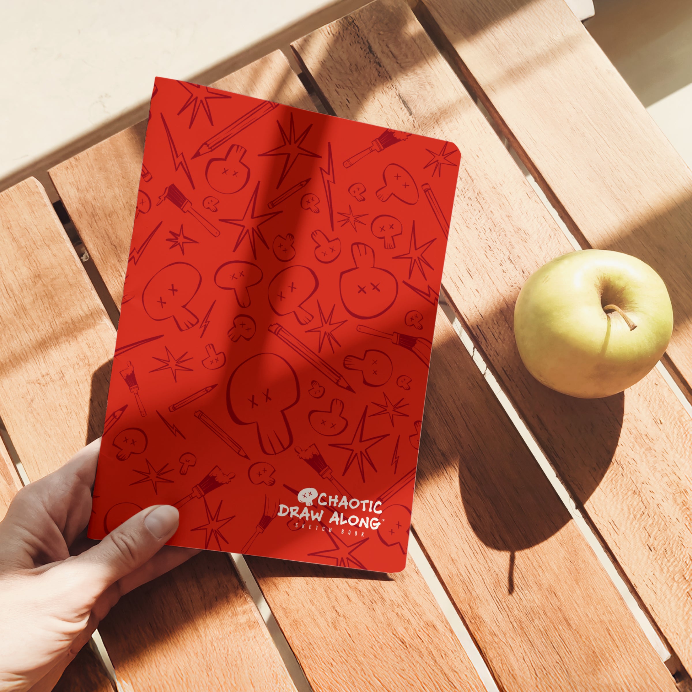 Person holding the red Chaotic Draw Along sketchbook on a wooden table with sunlight and an apple nearby.