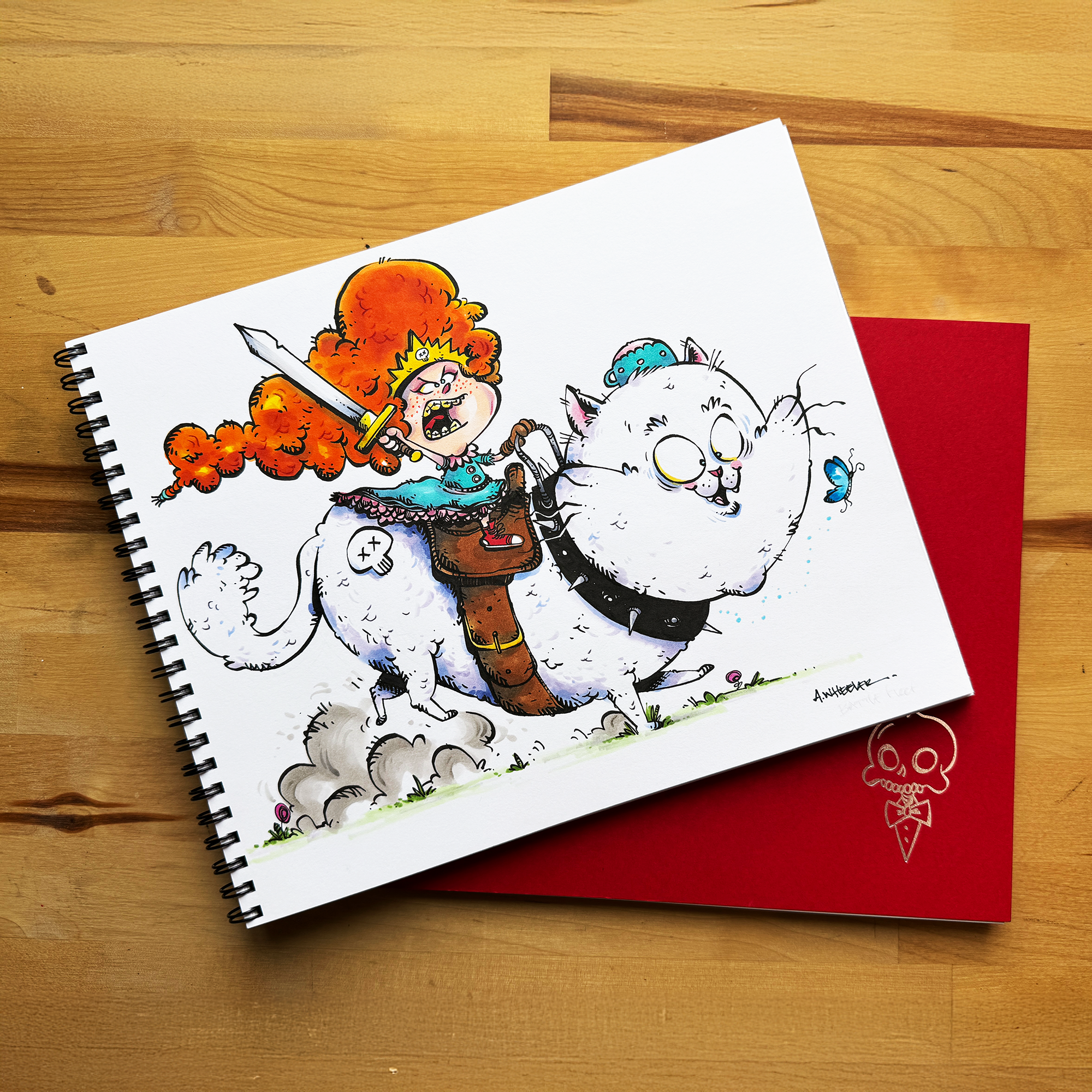 Spiral-bound sketchbook showing a colorful cartoon warrior riding a large white cat, placed on top of a red notebook on a wooden table.