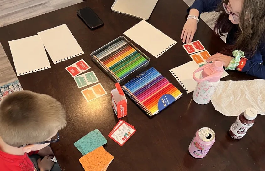 Two kids using Chaotic Draw Along prompt cards at a table with sketch pads, colored pencils, and drawing supplies.