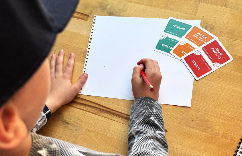 Child drawing at a table with Chaotic Draw Along prompt cards, sketchbook, and pencil for a screen-free family drawing activity.