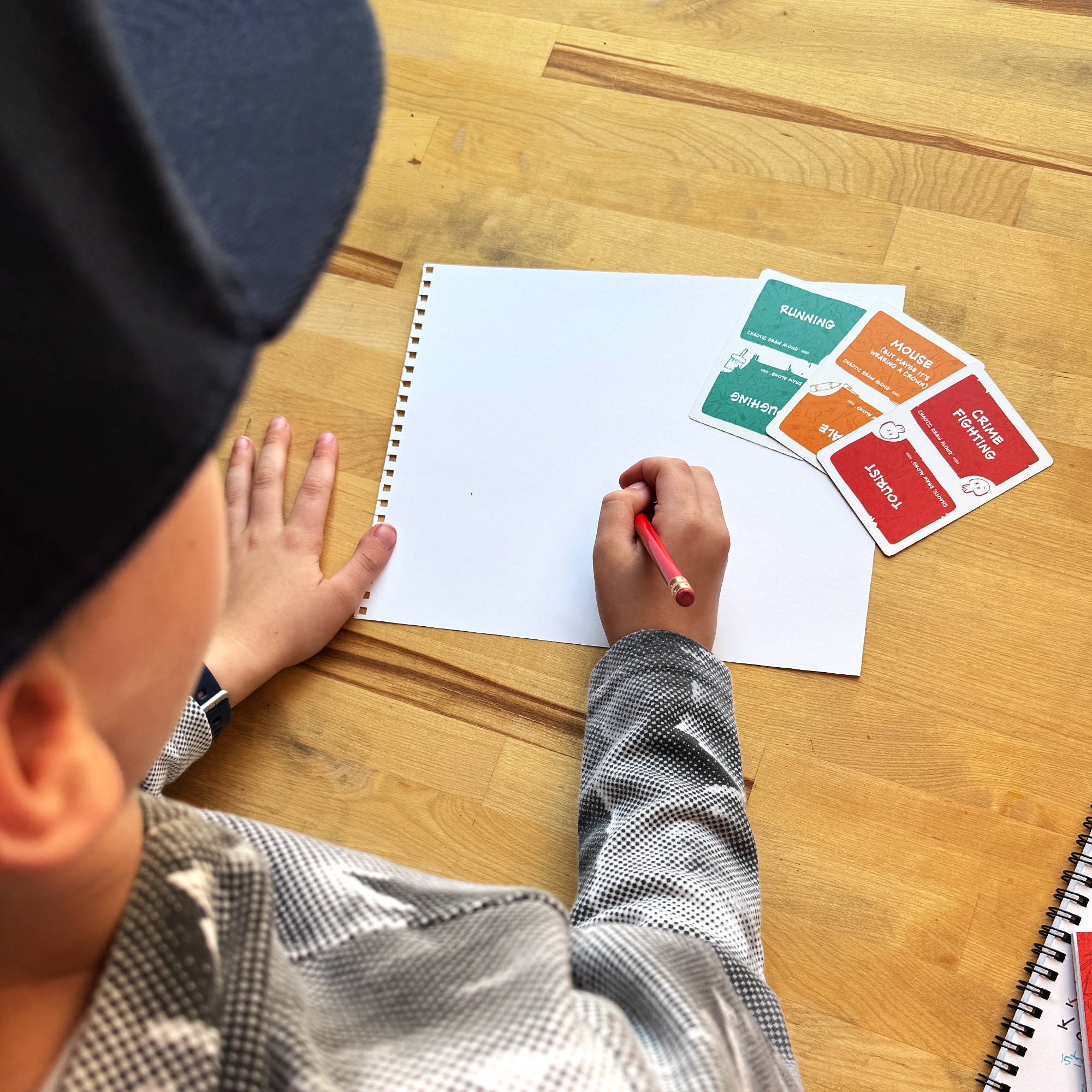 Child drawing with a red pencil on a blank sketchbook page, with Chaotic Draw Along prompt cards beside them on a wooden table.