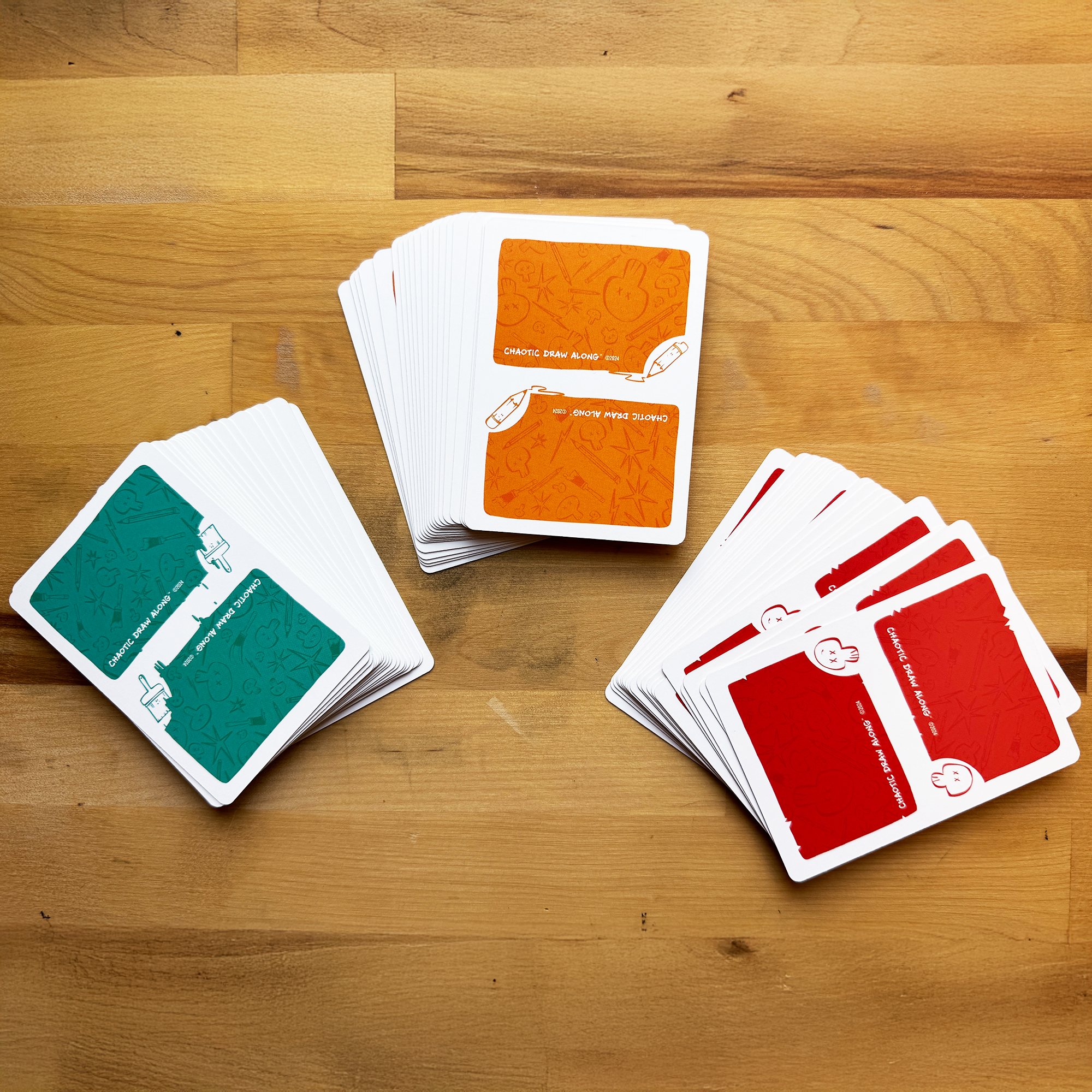 Three stacks of Chaotic Draw Along booster prompt cards in teal, orange, and red spread across a wooden table.
