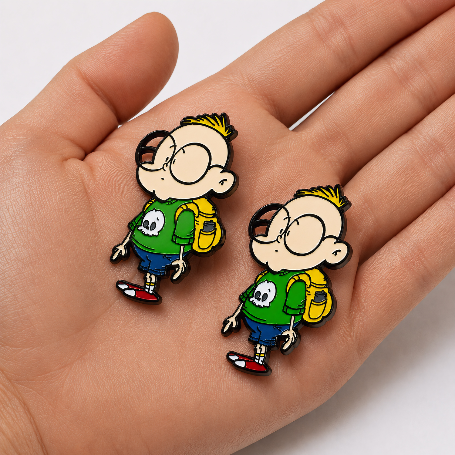 Two Gil hard enamel pins resting on a hand to show size and detail, featuring the Chaotic Draw Along character wearing a green shirt with a skull design and a yellow backpack.