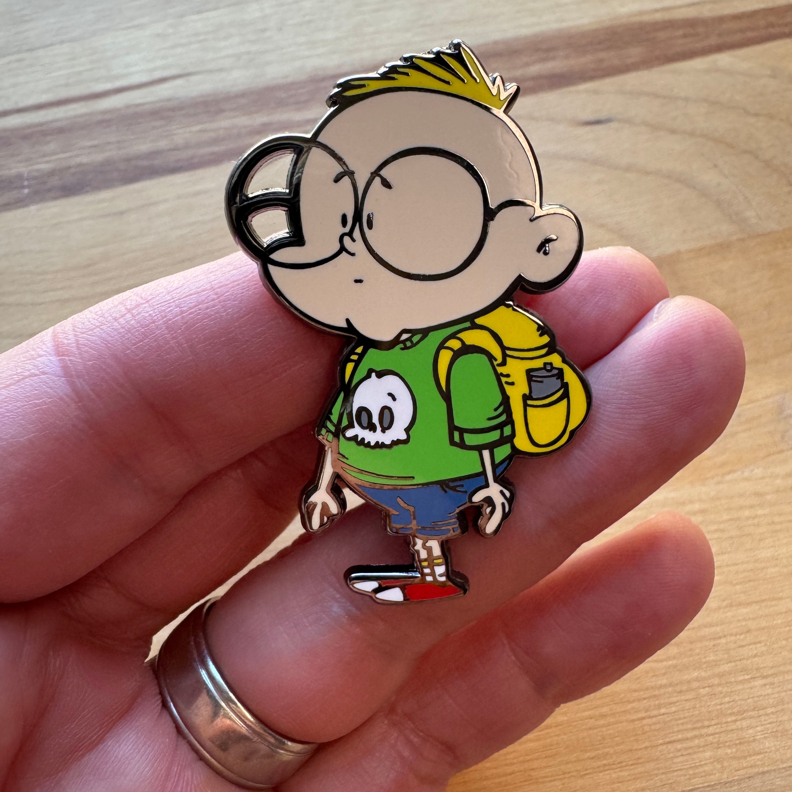 Close-up of the Gil hard enamel pin featuring the Chaotic Draw Along character wearing a green shirt with a skull design and a yellow backpack.