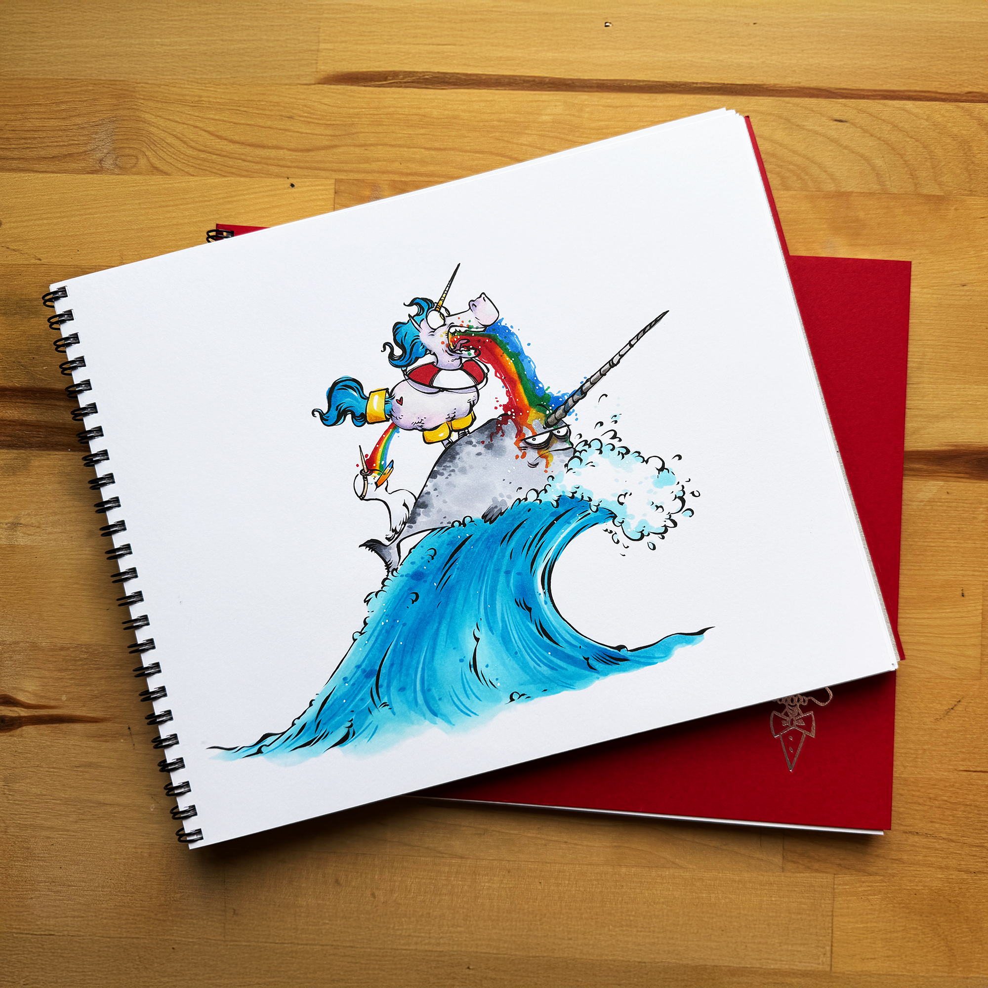 Spiral-bound sketchbook showing a colorful unicorn riding a narwhal through a blue wave, placed on a red notebook on a wooden table.