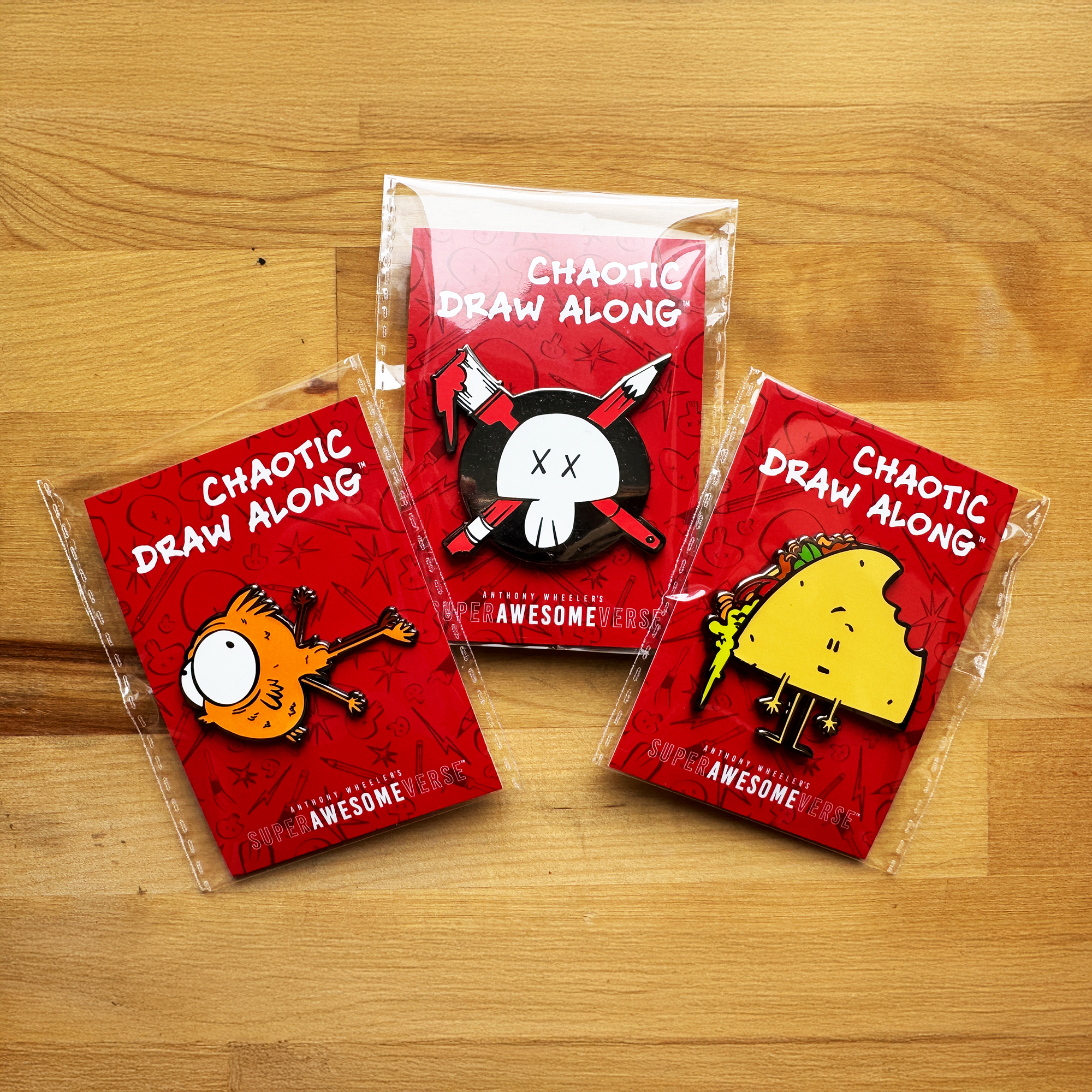 Three Chaotic Draw Along enamel pins in individual red packaging, featuring an orange character, skull logo, and taco character on a wooden table.