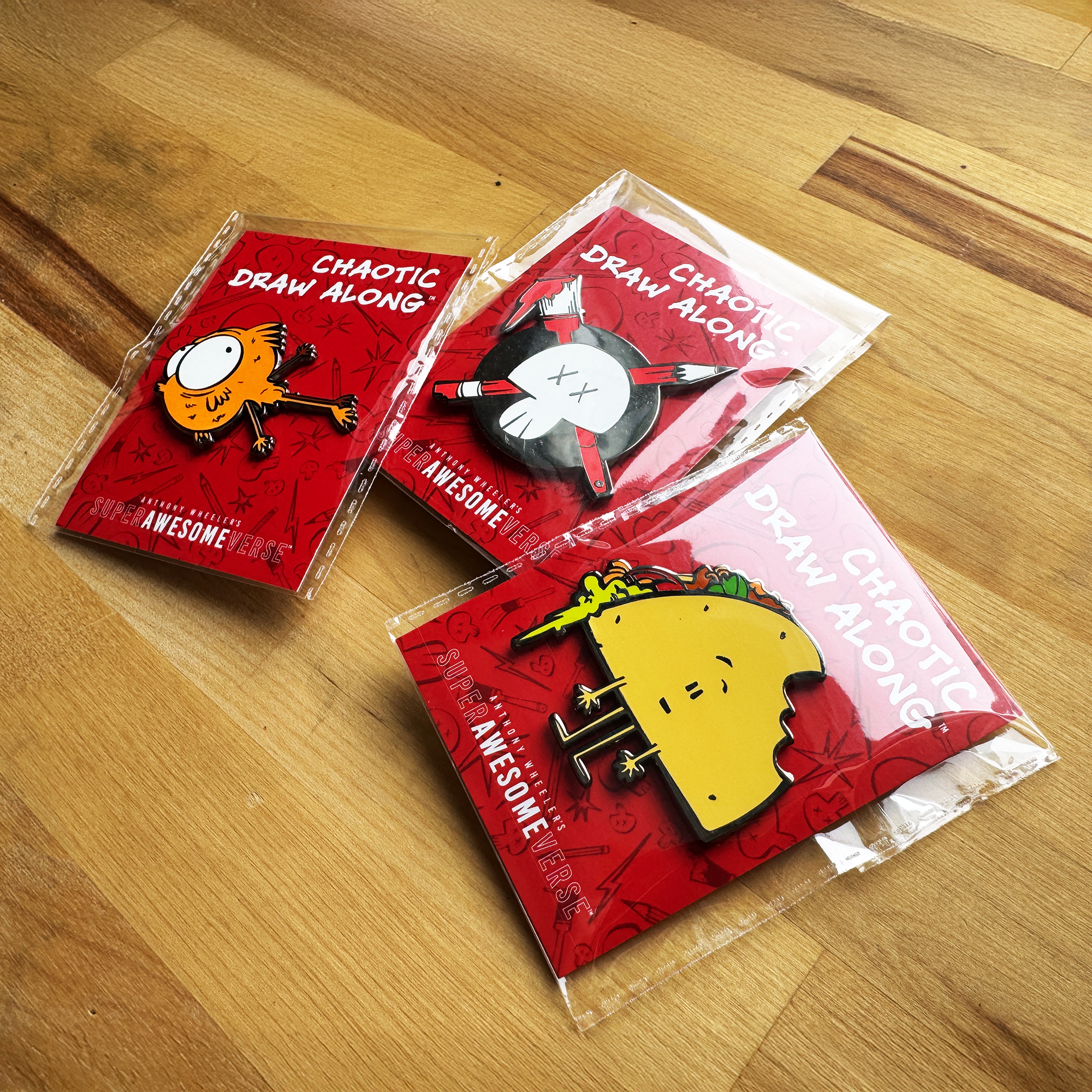 Three packaged Chaotic Draw Along enamel pins featuring an orange creature, skull logo, and taco character displayed on a wooden table.